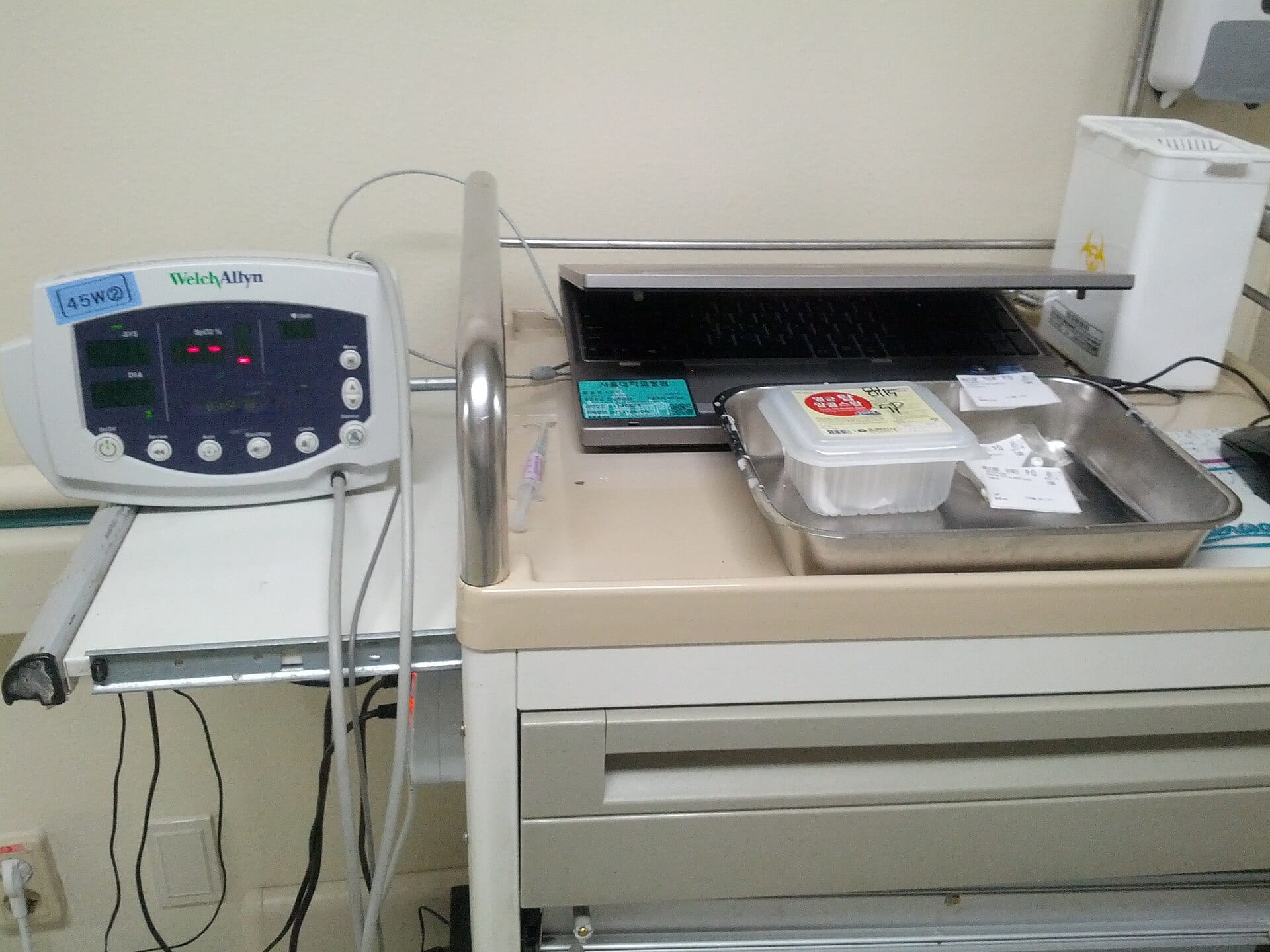 An automatic vital signs monitor on a cart beside a hospital bed, used for routine blood pressure checks on the ward