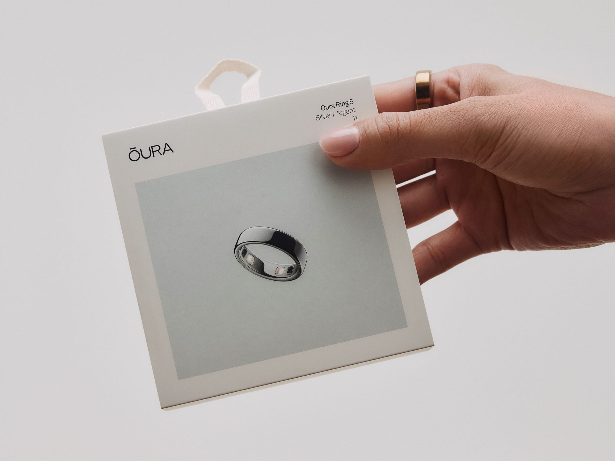 A hand holding the box of an Oura Ring 5, wearing a gold ring